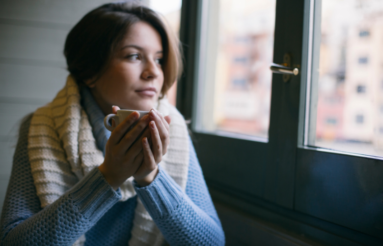 A woman holding a mug and wearing a scraf to keep warm as she looks out of the window.