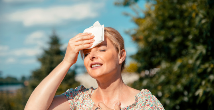 Picture of a lady standing in the sun holding a tissue to her head to prevent sweating.