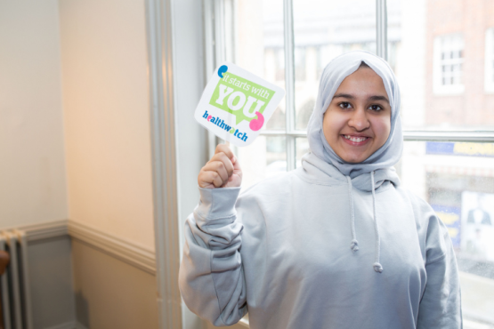 A volunteer holding up a small Healthwatch sign that says "It starts with you"